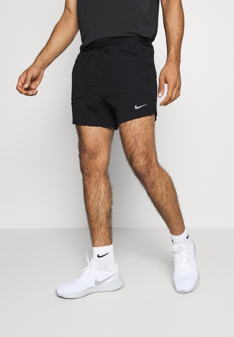 Homme portant un short noir Nike, des chaussettes blanches Nike et des chaussures de course blanches Nike, debout devant un fond clair uni.