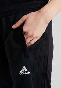Schwarze Adidas-Trainingshosen mit einer Reißverschlusstasche, die drei vertikale Streifen an den Seiten und ein weißes Logo am unteren linken Bein aufweisen.