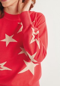 Rot gestrickter Pullover mit cremefarbenen Sternmustern, geripptem Halsausschnitt und Bündchen, weichem Material und lässiger Passform.
