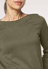 Maglione leggero a maniche lunghe verde oliva con una texture a costine. Presenta un ampio scollo e delle cuciture decorative sulla spalla.