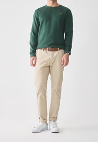 Felpa verde a maniche lunghe, chinos beige, cintura marrone e sneaker bianchi. Dettagli notevoli: logo semplice sul petto e orli dei pantaloni arrotolati.