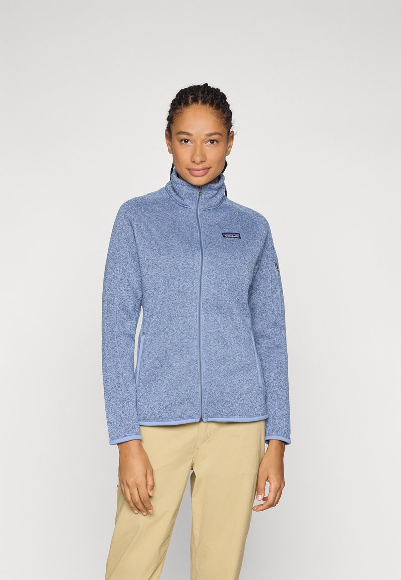 Giacca in pile azzurro chiaro con colletto alto, zip completa e una patch con logo sul petto. Texture liscia e design aderente.