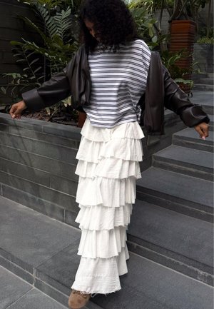 Ženska z kodrastimi lasmi, oblečena v črtasto srajco, belo večplastno maxi krilo in temno usnjeno jakno, stoji na kamnitih stopnicah blizu rastlin.