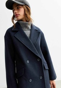 Cappotto doppiopetto in misto lana blu navy con revers, tasche laterali e grandi bottoni. Abbinato a un dolcevita grigio e un berretto.