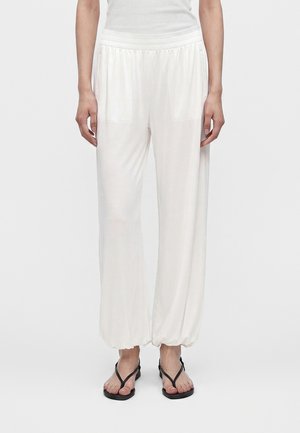Personne portant un pantalon blanc ample avec poignets et taille élastiques, des sandales noires et un haut blanc sans manches, debout devant un fond uni.