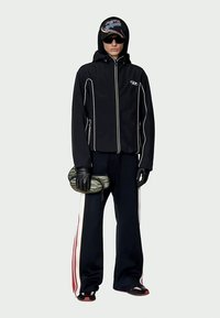 Person wearing black hooded jacket with white piping, black gloves, black pants with white and red side stripes, black sunglasses, and cap, holding striped pouch.