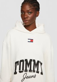Felpa bianca con cappuccio caratterizzata da una grande scritta nera "TOMMY" e "Jeans" in corsivo. Include un piccolo patch con logo nei colori navy, rosso e bianco. Tessuto morbido.