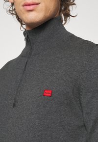 Maglione grigio con zip e colletto alto, realizzato in materiale testurizzato; presenta un logo quadrato rosso sul petto.