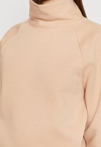 Sudadera de cuello alto color beige claro con una textura suave y lisa, mangas raglán y detalle de costuras mínimas a lo largo del cuello.