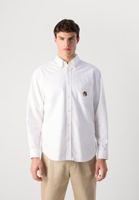 Jonge man die vooraan kijkt, gekleed in een wit overhemd met een geborduurd logo op de linker borstzak en beige broek tegen een effen achtergrond.