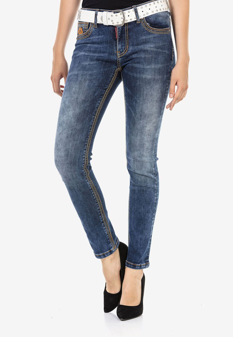 Schlanke blaue Denim-Jeans mit verwaschenen Stellen, orangen Nähten und einem weißen Nieten-Gürtel. Schwarze High heels vervollständigen das Outfit.