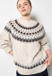 Beżowy sweter dzianinowy z okrągłym wykończeniem ozdobionym ciemnobrązowymi i niebieskimi wzorami. Odpinane ściągacze przy mankietach i u dołu, miękka faktura i luźny krój.