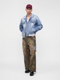 Jeune homme portant une casquette rouge, une veste en jean sur une chemise blanche, un pantalon cargo camouflage et des chaussures noires brillantes, debout les mains dans les poches.