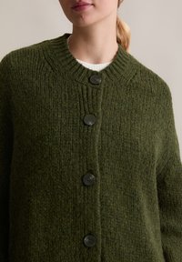 Cardigan en tricot vert avec une texture côtes, doté d'un col rond et de quatre boutons noirs à l'avant.