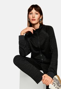 Schwarzer Trainingsanzug mit Reißverschluss und strukturierter Zebramusterung, dazu passende eng anliegende Hose und kontrastierende Bündchen; Modell trägt gemusterte Sneaker.