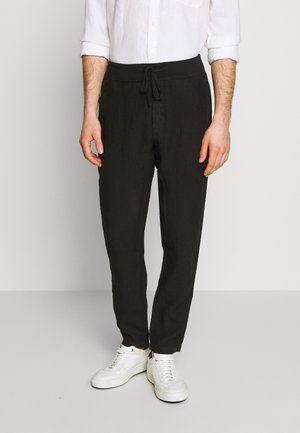 Mężczyzna w czarnych spodniach ze sznurkiem, białej koszuli z podwiniętymi rękawami oraz białych tenisówkach stoi na jednolitym tle.