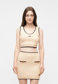 Νέα γυναίκα που φοράει μπεζ cropped τοπ με υφή και ταιριαστή μίνι φούστα με μαύρο περίγραμμα και μικρές διακοσμητικές τσέπες, στέκεται μπροστά σε ένα απλό φόντο.