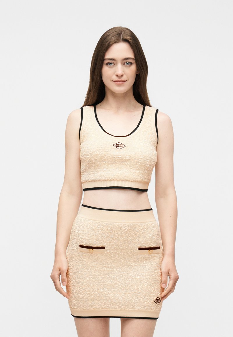 Νέα γυναίκα που φοράει μπεζ cropped τοπ με υφή και ταιριαστή μίνι φούστα με μαύρο περίγραμμα και μικρές διακοσμητικές τσέπες, στέκεται μπροστά σε ένα απλό φόντο.