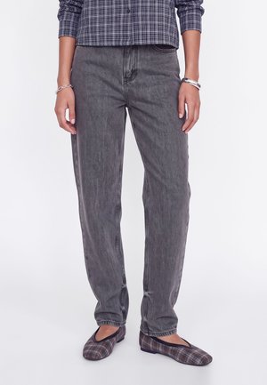 Personne portant un jean droit gris, une chemise à carreaux noir et blanc, des chaussures plates à carreaux et des bracelets argentés aux deux poignets.