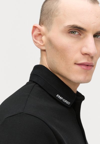 Black polo shirt with a textured collar featuring white "EMPORIO" lettering. Smooth fabric with a fitted design, highlighting the neckline.