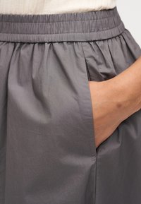 Χέρι που ξεκουράζεται μέσα στην πλαϊνή τσέπη γκρι χαλαρών παντελονιών με ελαστική ζώνη μέσης και μπεζ τοπ.