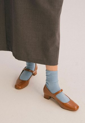 Bruine Mary Jane schoenen met lage hak, gedragen met lichtblauwe sokken en een lange donkerbruine rok, staande op een lichte vloer.