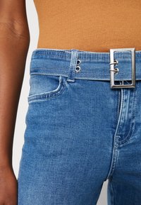 Primer plano de una persona con jeans de mezclilla azules, un cinturón a juego y una blusa marrón con canalé.