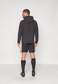Black athletic hoodie with a textured pattern, worn over black shorts featuring yellow side stripes. Black long socks and cleats visible.