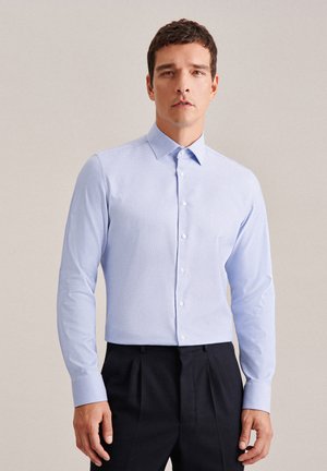 Camicia azzurra a maniche lunghe con colletto a button-down, tessuto texturizzato, sei bottoni sul fronte, indossata con pantaloni scuri.