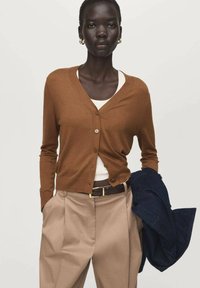 Bruine gebreide cardigan met V-hals, knoopsluiting, gedragen over een wit shirt. Gecombineerd met beige broek en een donkerblauwe jas in de hand.