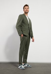 Oliven grøn jakkesæt med en tætsiddende blazer og slanke bukser, parret med en hvid T-shirt og grønne/hvide sneakers. Glat tekstur, moderne design.