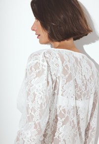 Weißes Spitzenoberteil mit floralen Mustern, transparenter Textur und lockerer Passform. Das Design umfasst einen weiten Halsausschnitt und lange Ärmel.