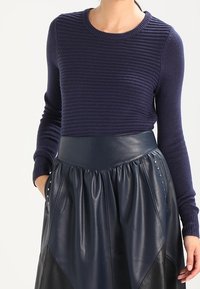 Een vrouw in een marineblauwe ribgebreide trui, gestoken in een hoge taille marineblauwe leren rok met zijzakken en studs.