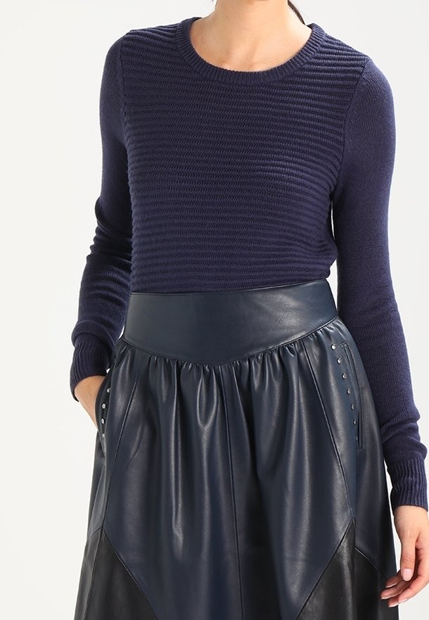 Een vrouw in een marineblauwe ribgebreide trui, gestoken in een hoge taille marineblauwe leren rok met zijzakken en studs.