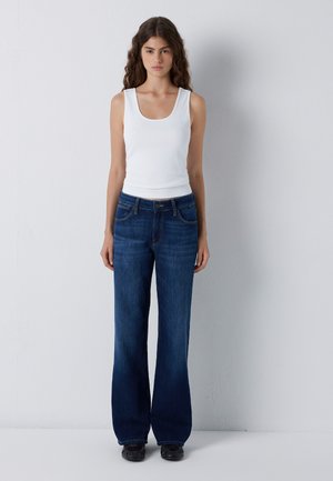 Young woman with long curly hair wearing a white sleeveless top, blue wide-leg jeans, and black shoes standing against a plain wall.