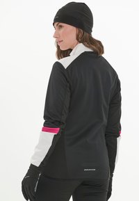 Schwarze Sportjacke mit pinken und grauen Akzenten, strukturierter Stoff, hoher Kragen und ergonomische Passform. Text-Logo am unteren Saum.