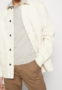Jasnobeżowa kurtka z czarnymi guzikami, pojedynczą kieszenią na piersi, noszona na jasnoszarej koszuli i brązowych spodniach. Bawełniana tkanina, casualowy design.