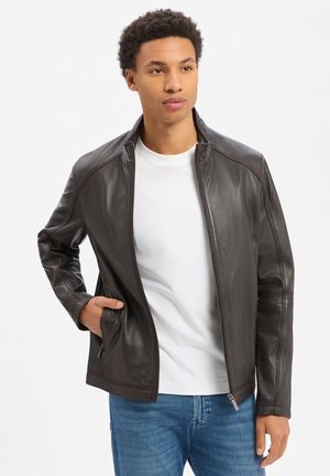 Junger Mann, der eine dunkelbraune Lederjacke über einem weißen T-Shirt und blauen Jeans trägt und mit einer Hand in der Tasche steht.