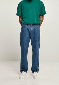 Camisa verde de manga corta con cremallera combinada con jeans de mezclilla azul. Los jeans tienen un corte recto y un diseño clásico de cinco bolsillos. Zapatillas blancas completan el conjunto.