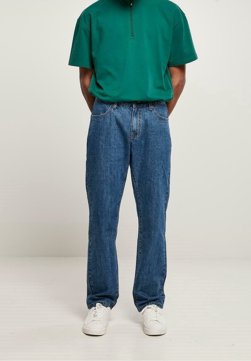 Camisa verde de manga corta con cremallera combinada con jeans de mezclilla azul. Los jeans tienen un corte recto y un diseño clásico de cinco bolsillos. Zapatillas blancas completan el conjunto.