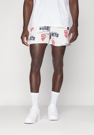 Muž oblečený v bílých kraťasech s potiskem San Francisco Giants, bílých teniskách a bílých ponožkách po lýtka, stojící před jednobarevným pozadím.