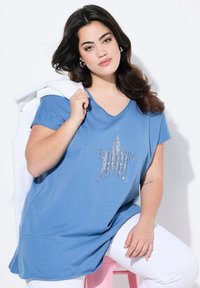 Túnica corta de manga corta azul con escote en V y patrón de estrellas plateadas en el centro. Tela suave, corte relajado, combinada con pantalones blancos.