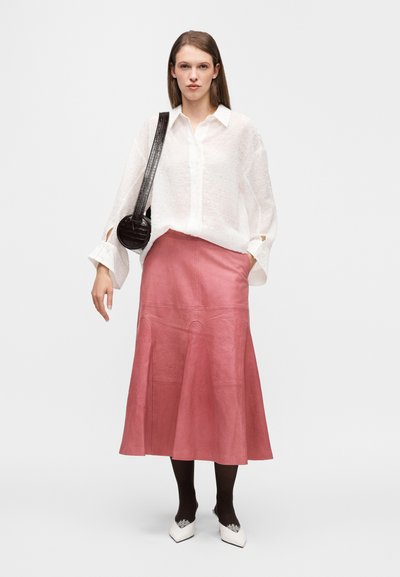 Chemise blanche texturée à coupe ample avec manches larges, associée à une jupe midi en cuir rose. Sac noir et chaussures blanches à bout pointu.