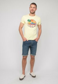 T-shirt jaune clair avec un graphique coloré "Miami", associé à un short en denim roulé et des chaussures légères à enfiler. Tenue décontractée d'été.