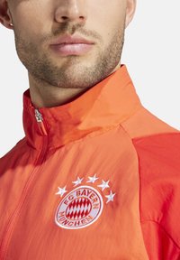 Chaqueta naranja de cremallera con cuello alto, que presenta un logo bordado del FC Bayern München y estrellas, fabricada con un material ligero y suave.