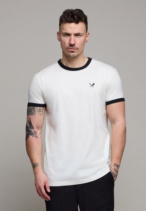 Mann med kort mørkt hår iført hvit t-skjorte med svart kant og logo med kryssede padler, som viser tatoveringer på begge armer mot ensfarget bakgrunn.