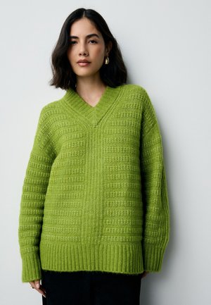 Kvinde med skulderlangt mørkt hår iført guldbøjleøreringe og en struktureret limegrøn V-hals sweater, stående foran en hvid væg.