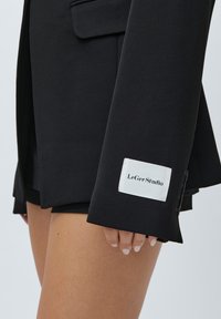 Prim-plan al unei persoane îmbrăcate cu un sacou negru croiat, având o etichetă albă "LeGer Studio" pe mânecă, asortat cu pantaloni scurți negri.