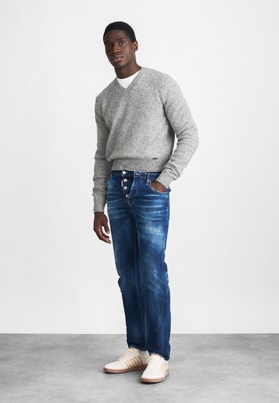 Grijze gestructureerde trui, blauwe denim jeans met knopenrij, en witte sneakers met beige accenten. Staande figuur tegen een effen achtergrond.