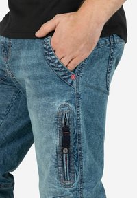 Blaue Denim-Hosen weisen ein leicht verwaschenes Design auf, mit einer seitlichen Reißverschlusstasche und kontrastierendem Stich. Der Stoff hat eine strukturierte Oberfläche.